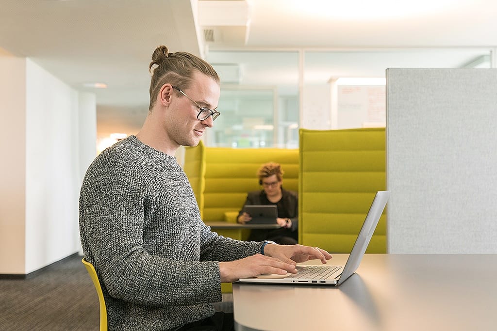Ein junger Mann sitzt an einem Tisch und arbeitet an einem Laptop. Im Hintergrund ist eine arbeitende Frau zu sehen.